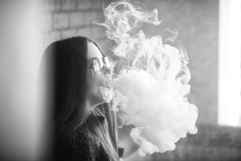 Vape teenager with  problem skin. Portrait of young cute girl in sunglasses smoking an electronic cigarette in the bar. Bad habit. Black nad white.